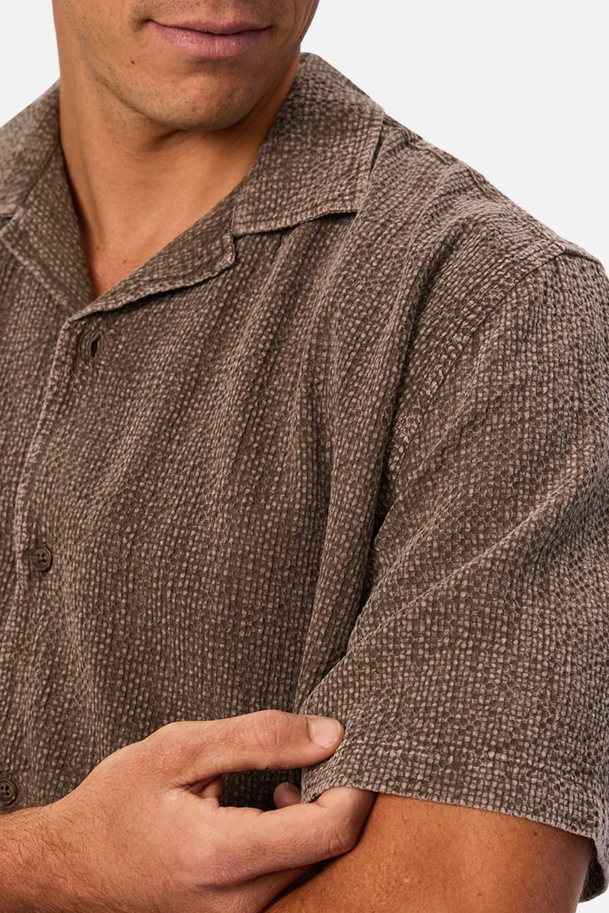 A man is shown from the shoulders up wearing the Industrie The Canyon Short Sleeve Shirt, a lightweight camp-collar shirt, gently pinching its textured sleeve with his right hand.
