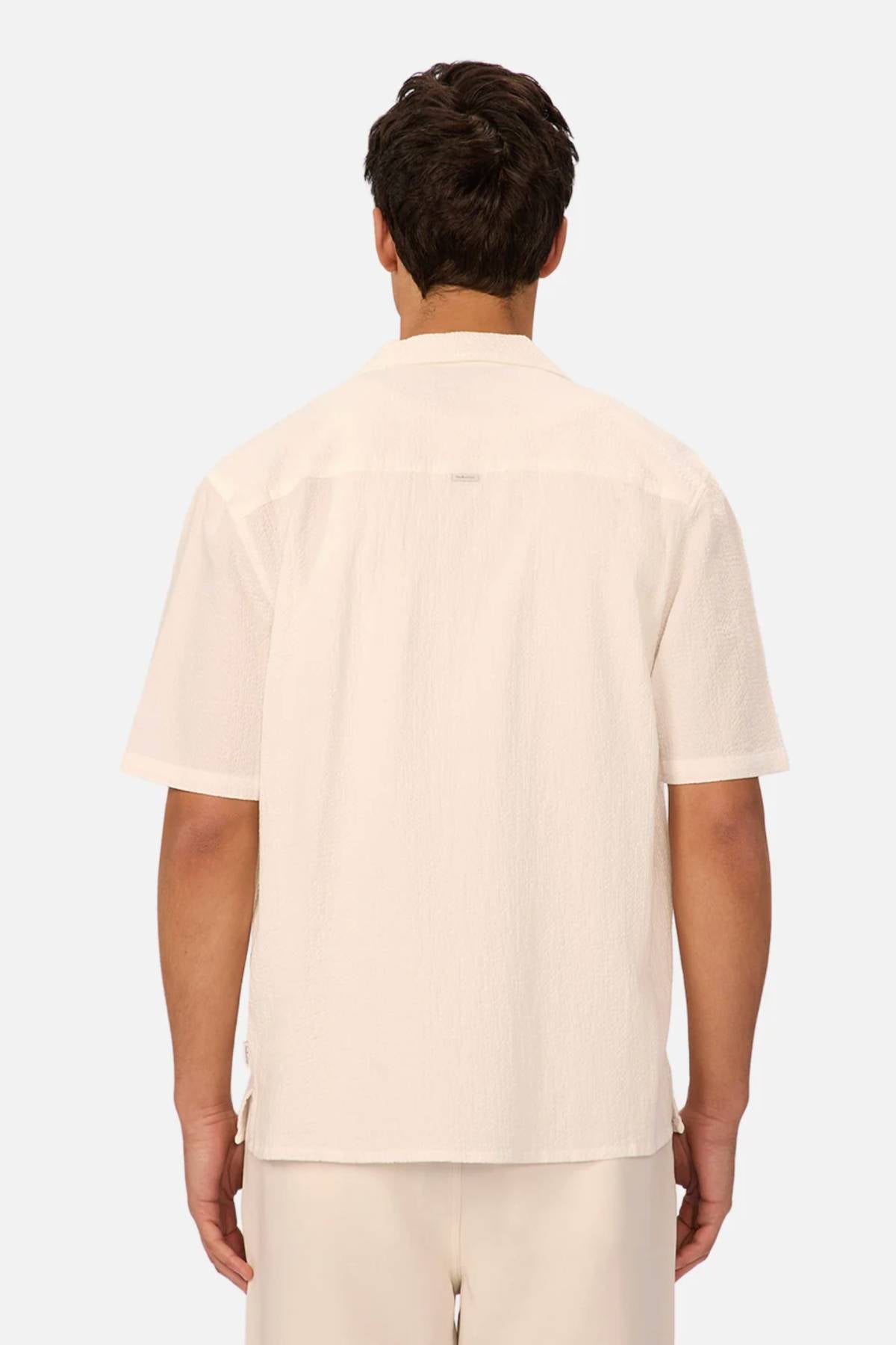 A man is seen from the back wearing the Industrie The Canyon Short Sleeve Shirt, a light-colored camp-collar short sleeve shirt, with matching pants against a plain light gray background.