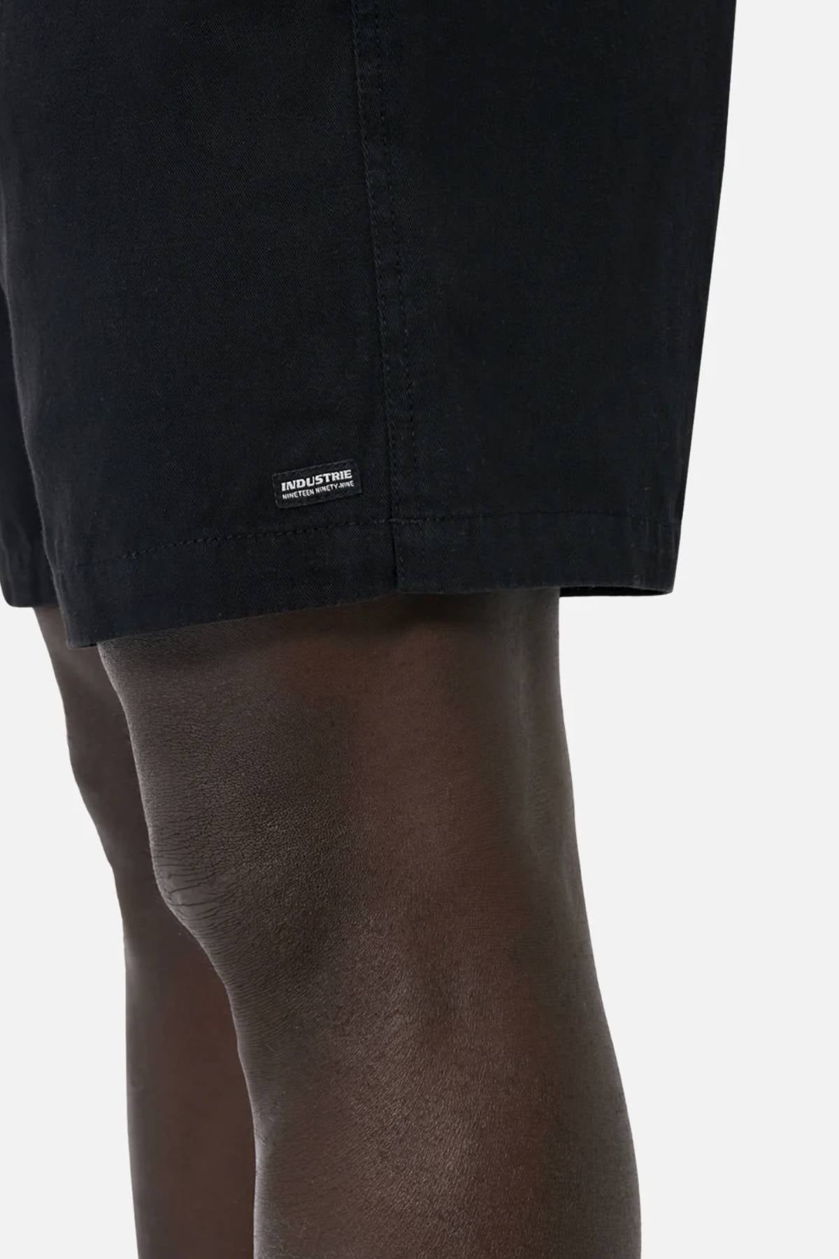 A close-up shows a person wearing Industrie The Dublin Short, black men's summer cotton-linen shorts. The lower part displays a small rectangular label, with part of the thigh and knee visible against a plain light background.
