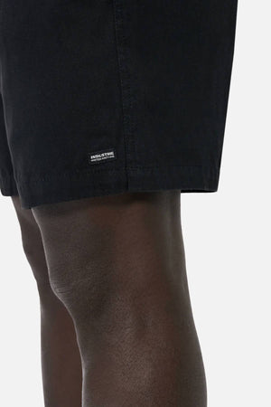 A close-up shows a person wearing Industrie The Dublin Short, black men's summer cotton-linen shorts. The lower part displays a small rectangular label, with part of the thigh and knee visible against a plain light background.