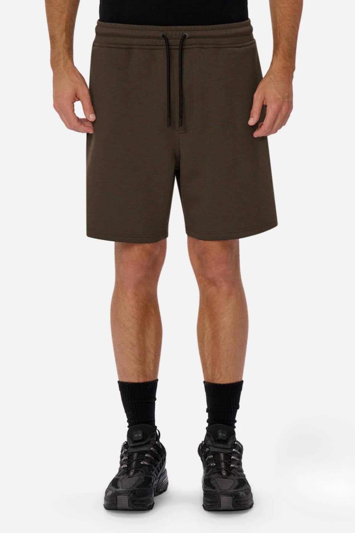 A person wears the Industrie The Tech Track Short in olive green with a black shirt, black socks, and black sneakers, standing against a plain white background to showcase the shorts' comfort and versatile style.