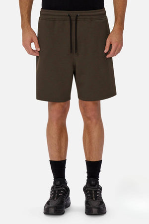 A person wears the Industrie The Tech Track Short in olive green with a black shirt, black socks, and black sneakers, standing against a plain white background to showcase the shorts' comfort and versatile style.