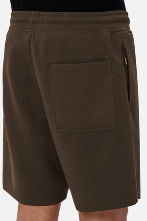 A close-up of a person wearing Industrie The Tech Track Short in dark olive green, featuring an elastic waistband, side zipper pocket, and back patch pocket. The comfortable design is paired with a black shirt, showing the person’s legs.