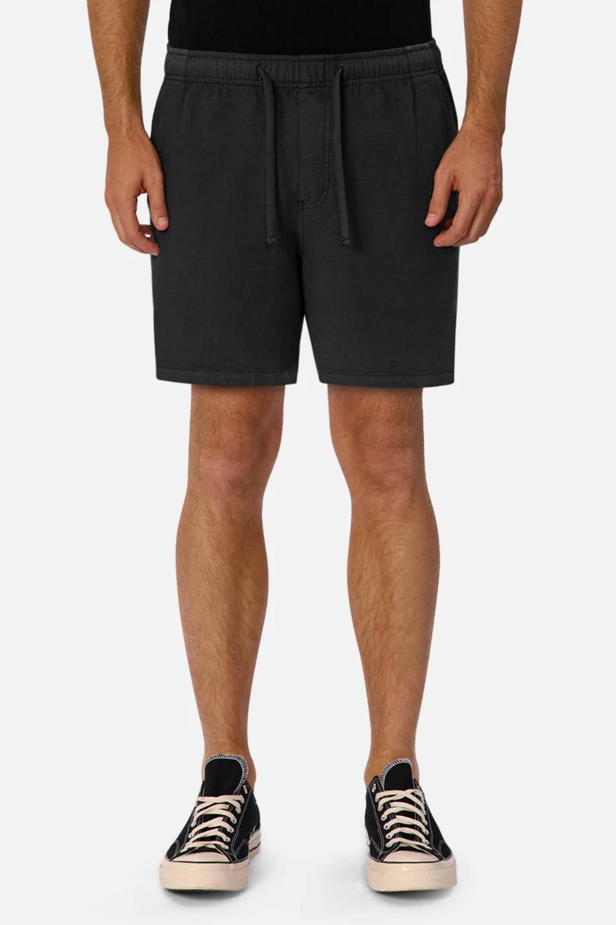 Wearing Industrie The Mallorca Short in black and black low-top sneakers with white soles, the lower half of a person is shown standing against a plain white background.
