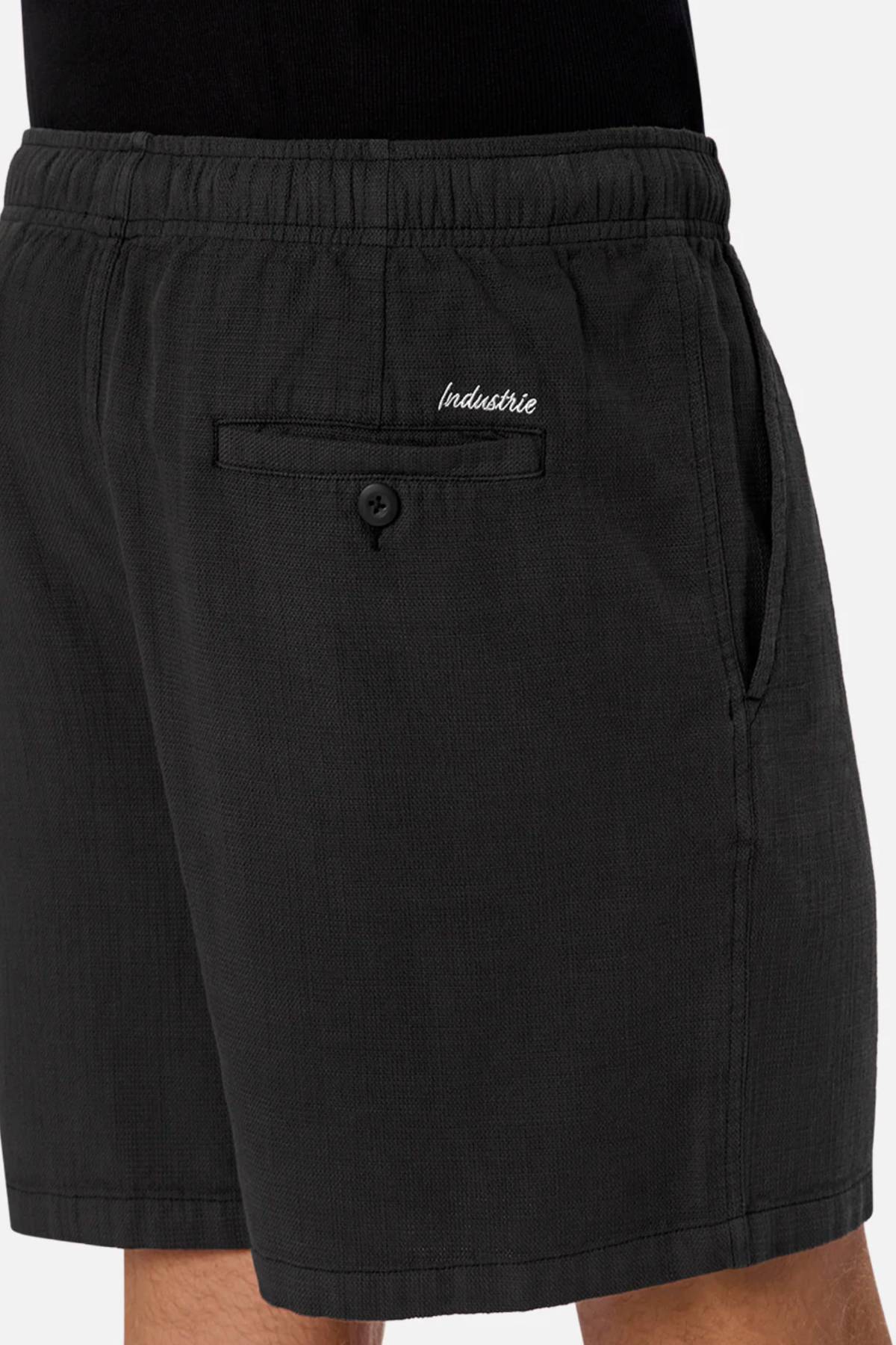 A close-up shows a person wearing Industrie The Mallorca Short in black cotton, featuring an elastic waistband, side pocket, buttoned back pocket with "Industrie" embroidered above it, paired with a black top.