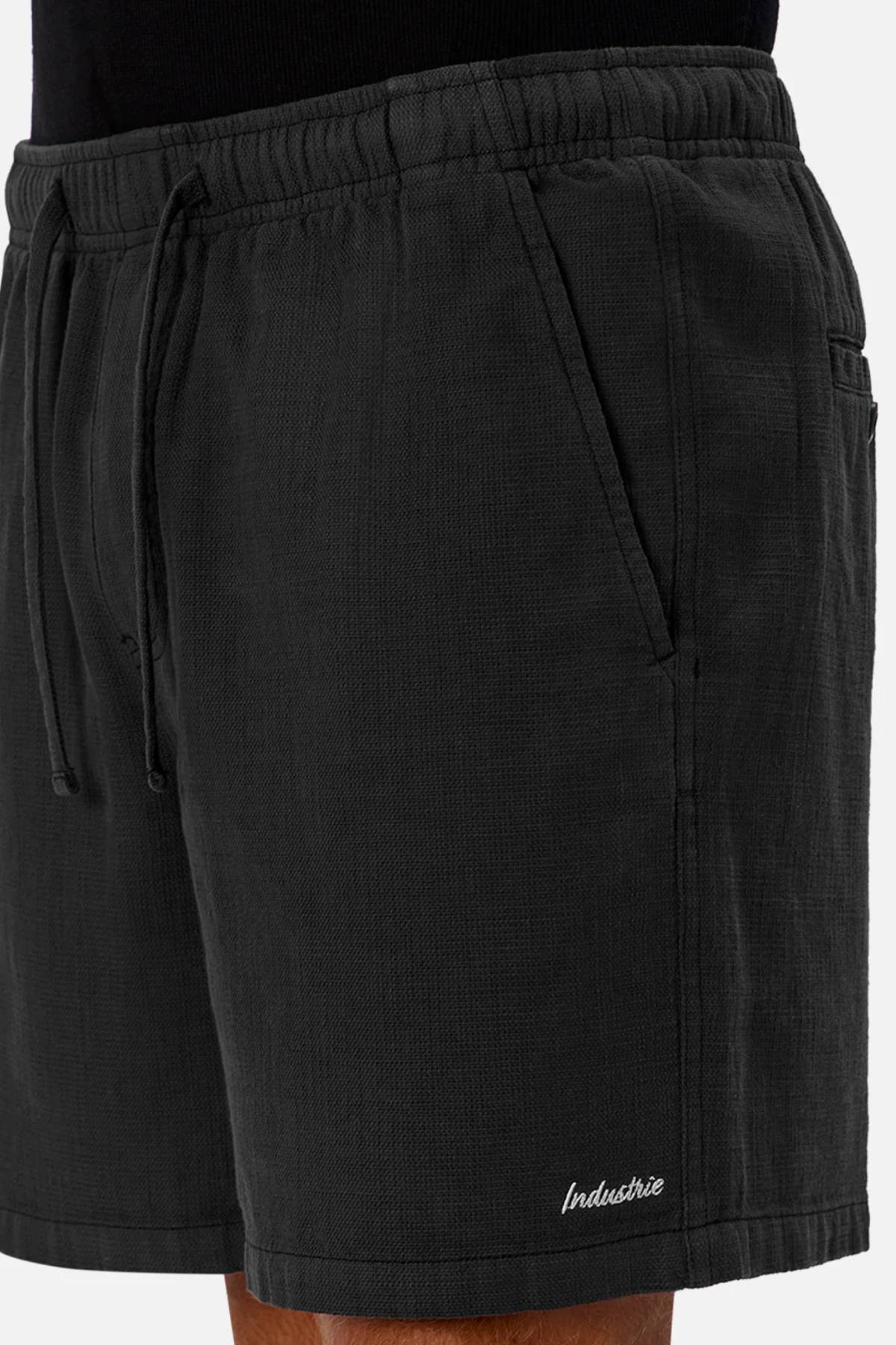 A close-up of a person wearing Industrie The Mallorca Short—men’s cotton shorts with pockets, textured fabric, and “Industrie” embroidered near the hem—paired with a black top.
