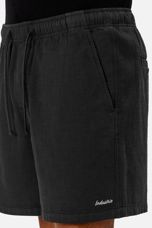 A close-up of a person wearing Industrie The Mallorca Short—men’s cotton shorts with pockets, textured fabric, and “Industrie” embroidered near the hem—paired with a black top.