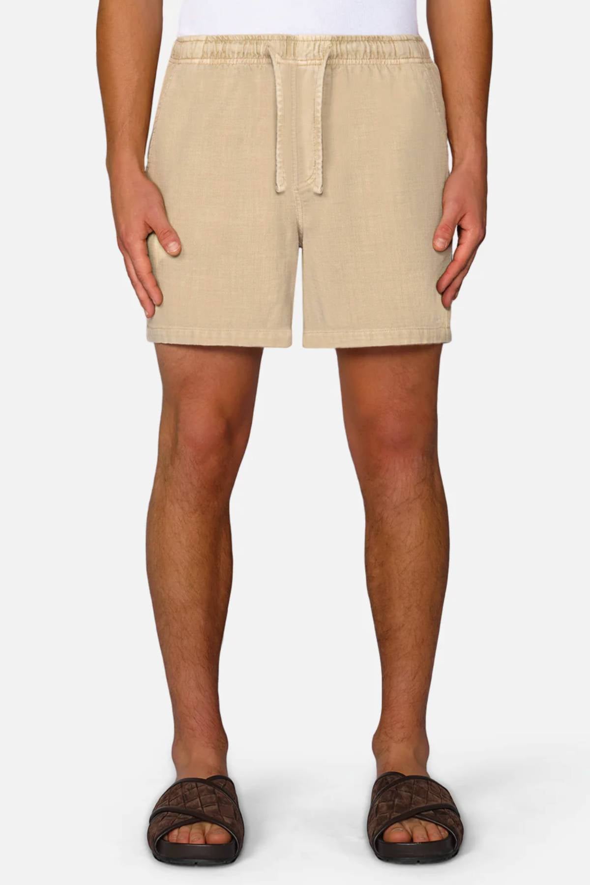 A person wears Industrie The Mallorca Short in beige, a white shirt, and dark brown slide sandals, standing facing forward against a plain background.