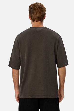 A person with short, light brown hair is seen from the back wearing a relaxed fit Industrie The Marlon Tee in dark gray and black pants against a plain light background.