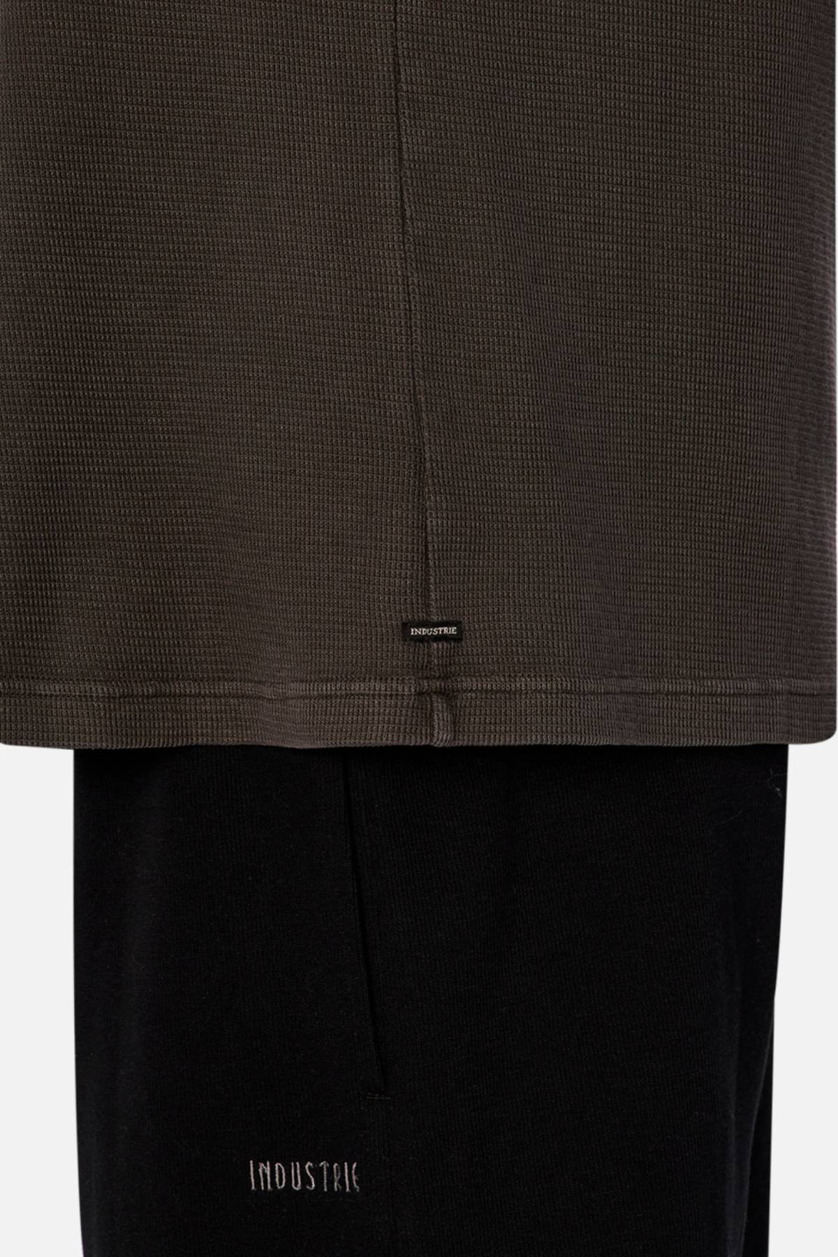 A close-up of the lower part of a relaxed fit Industrie The Marlon Tee in dark waffle knit fabric worn over black pants, both with subtle "INDUSTRIE" logos near the hems, set against a plain white background.