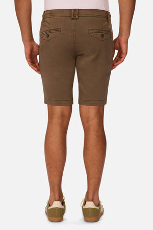 A person is shown from the back wearing Industrie The New Drifter Cuba Short—brown, slim-fit men's shorts with buttoned back pockets—a light pink shirt tucked in, and olive green sneakers with white soles.