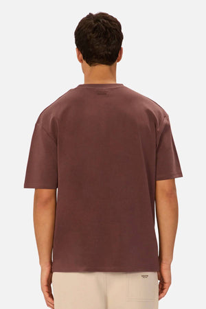 A person with short dark hair is shown from the back, wearing the Industrie The Newport Tee in dark brown with light beige pants against a plain white background.