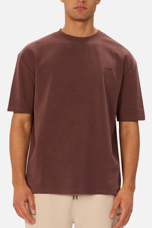 A man wears the Industrie The Newport Tee in brown—an oversized, loose-fitting shirt with small chest text—paired with light beige pants, standing against a plain white background.