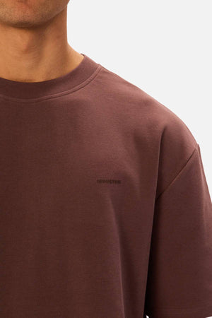 A person wearing the Industrie The Newport Tee in brown, an oversized crew-neck style with a subtle logo on the left chest. The image is cropped at the chin, highlighting the shoulder and torso against a plain background.