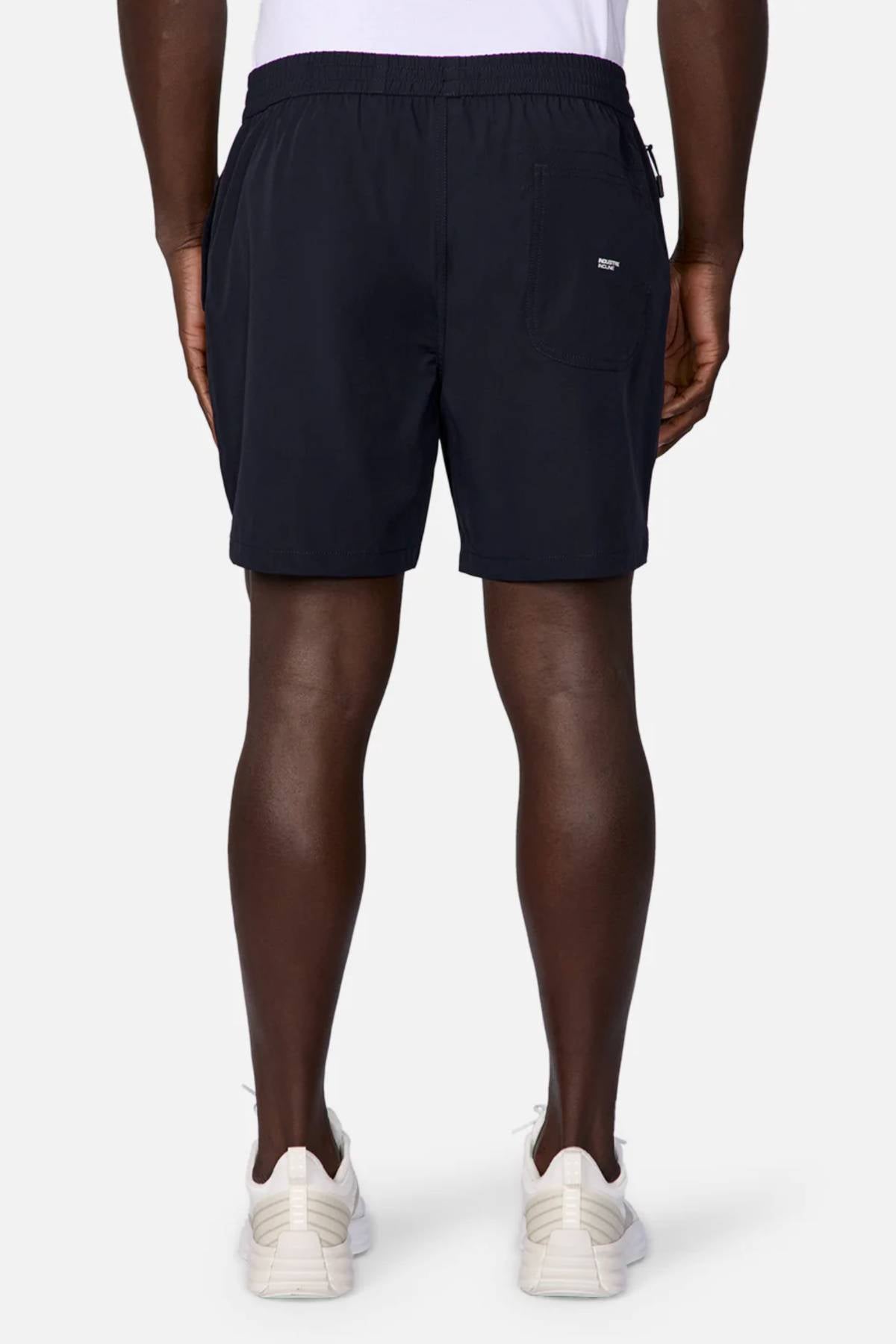 A person stands facing away in white sneakers and Industrie The Owens Short—black, quick-drying, above-knee shorts with an elastic waistband and back pocket—paired with a white shirt, set against a plain, light background.