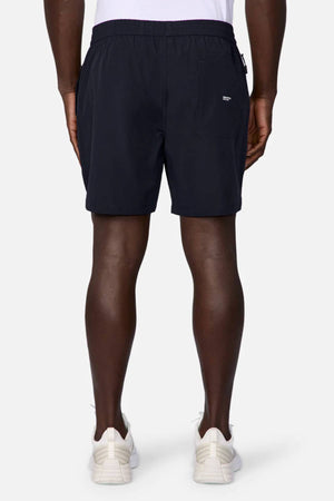 A person stands facing away in white sneakers and Industrie The Owens Short—black, quick-drying, above-knee shorts with an elastic waistband and back pocket—paired with a white shirt, set against a plain, light background.