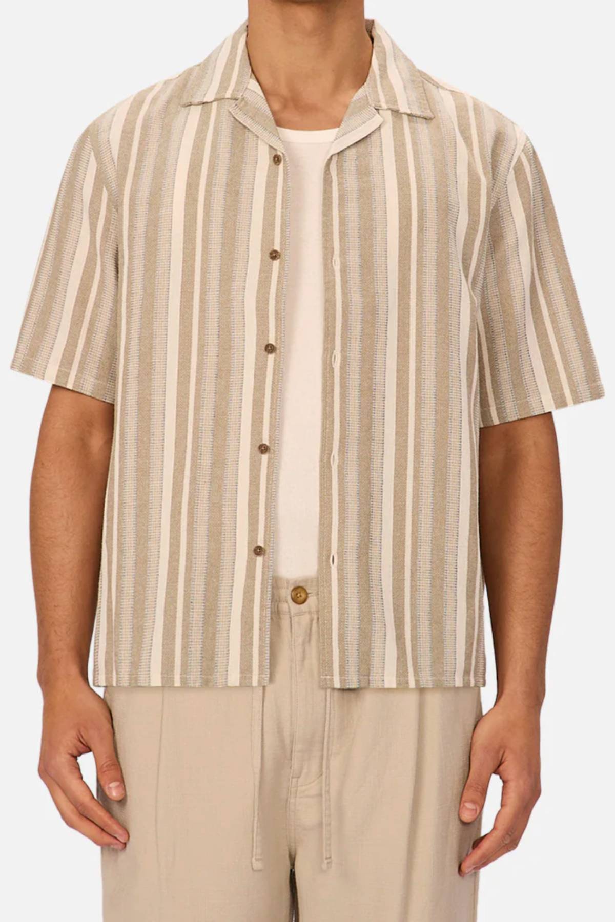 A man wears the Industrie The Textured Livorno Short Sleeve Shirt—beige and white vertical stripes, relaxed fit, short sleeves, camp collar—over a white T-shirt and light beige pants. Only his torso and arms are visible.