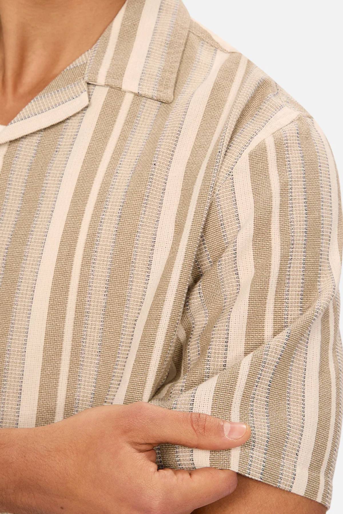 A person wears the Industrie The Textured Livorno Short Sleeve Shirt, featuring a relaxed fit and light blue-beige vertical stripes with a camp collar, holding the sleeve. Only the upper torso and arm appear against a plain background.