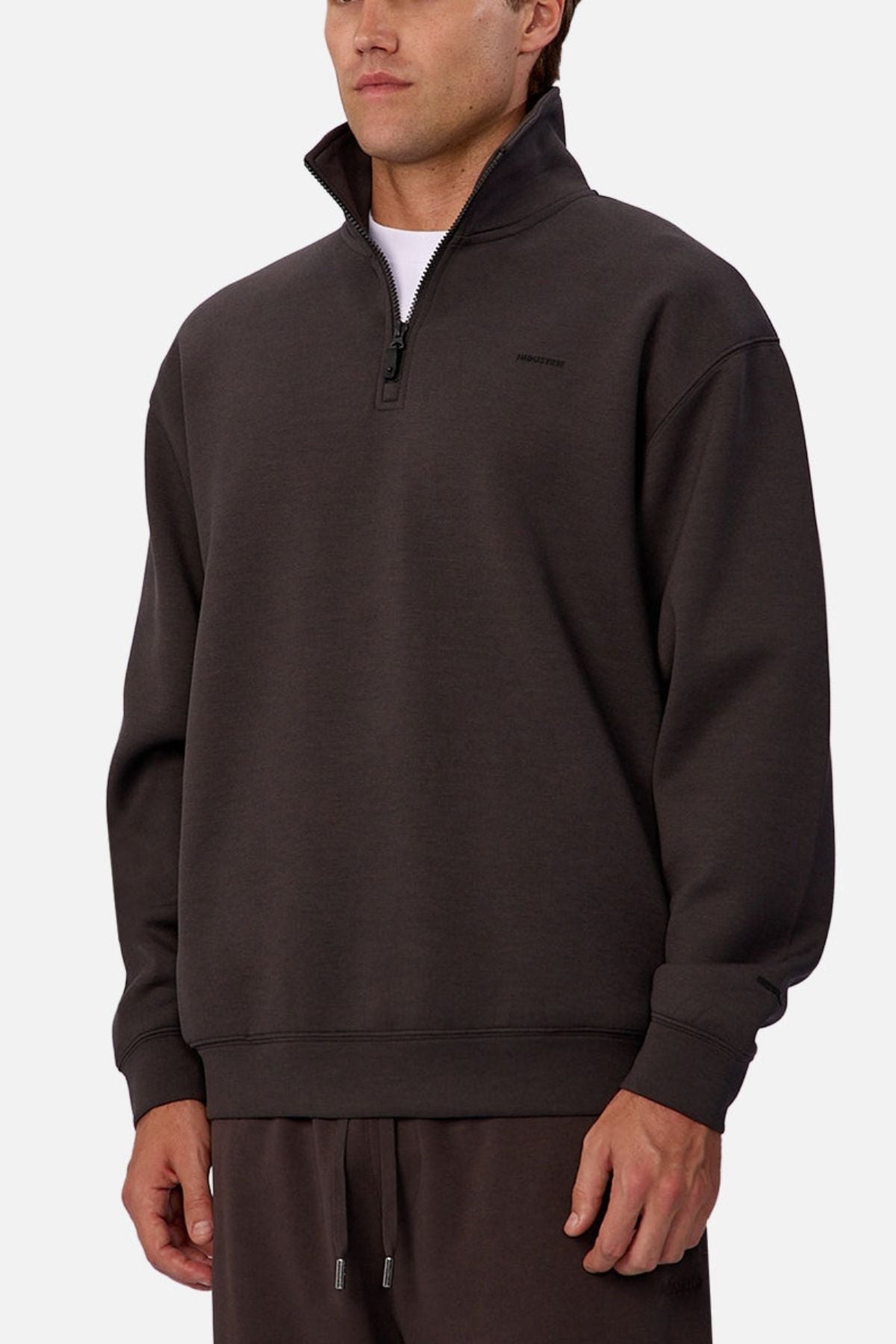 A man wears the Industrie The Tech Staunton Track Top, a dark brown athletic pullover with a high collar, paired with matching drawstring pants, standing against a plain white background.