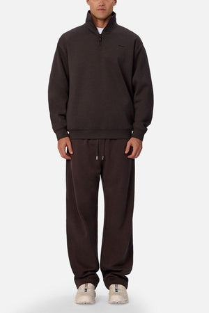 A person faces forward, dressed in the Industrie The Tech Staunton Track Top—a loose-fitting dark brown athletic pullover—with matching wide-leg sweatpants and white sneakers, standing against a plain light background.