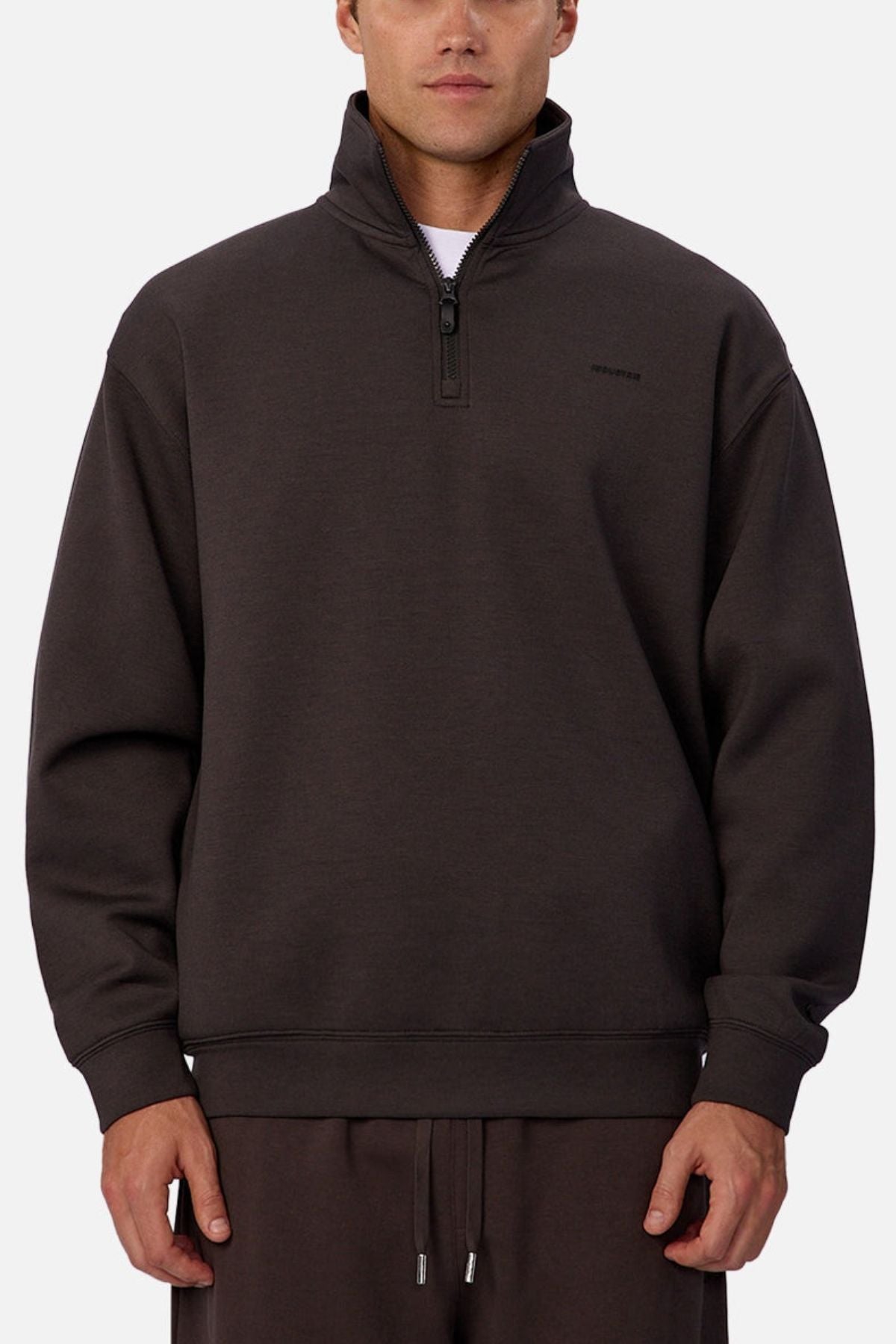 A person wears the Industrie The Tech Staunton Track Top in dark brown with a high collar and subtle left chest text, paired with matching drawstring pants, standing against a plain light background.