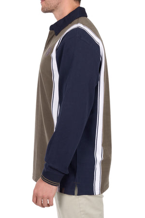 A man is seen from the side wearing the Thomas Cook Travis Rugby, a long-sleeve shirt with navy blue, olive green, and white vertical panels, paired with light-colored pants. His face is partially out of frame.