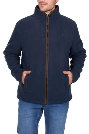 A man wearing a navy blue Thomas Cook Adrian Jacket with brown trim and blue jeans stands facing forward, hands in his jacket pockets, against a white background.