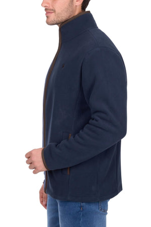A man stands in profile, wearing the Thomas Cook Adrian Jacket—a navy blue heavyweight polar fleece with brown trim, stand-up collar, and side pockets—paired with blue jeans against a white background.