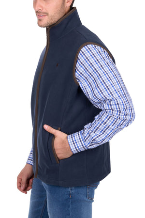 A man stands against a white background wearing the Thomas Cook Adrian Vest over a blue and white plaid shirt with blue jeans, one hand in his vest pocket.