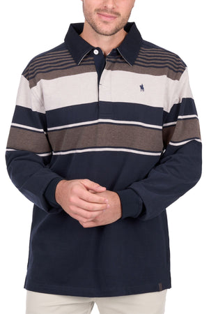 A man stands with his hands clasped, wearing the Thomas Cook Bryan Stripe Rugby—a navy, beige, and brown striped long-sleeve polo shirt with a collar—paired with light pants.