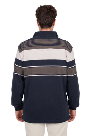 A man with short dark hair and light pants stands with his back to the camera, wearing the Thomas Cook Bryan Stripe Rugby—a long-sleeve, collared shirt with horizontal navy blue, gray, and white stripes.