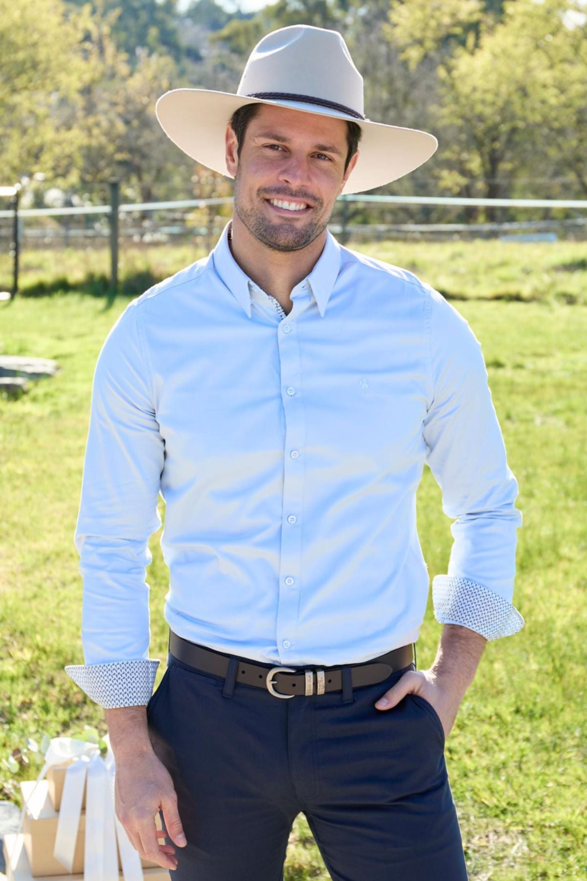 A man in a light blue Thomas Cook Callum Tailored Shirt and dark pants smiles outdoors on a sunny day, standing on grass with trees and a fence behind him.