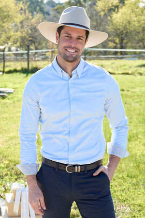 A man in a light blue Thomas Cook Callum Tailored Shirt and dark pants smiles outdoors on a sunny day, standing on grass with trees and a fence behind him.