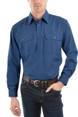 A man wearing the Thomas Cook Heavy Drill Half Placket Shirt, a long-sleeve blue work shirt with two chest pockets, paired with dark jeans and a black belt, stands against a white background. His face is partially out of frame.