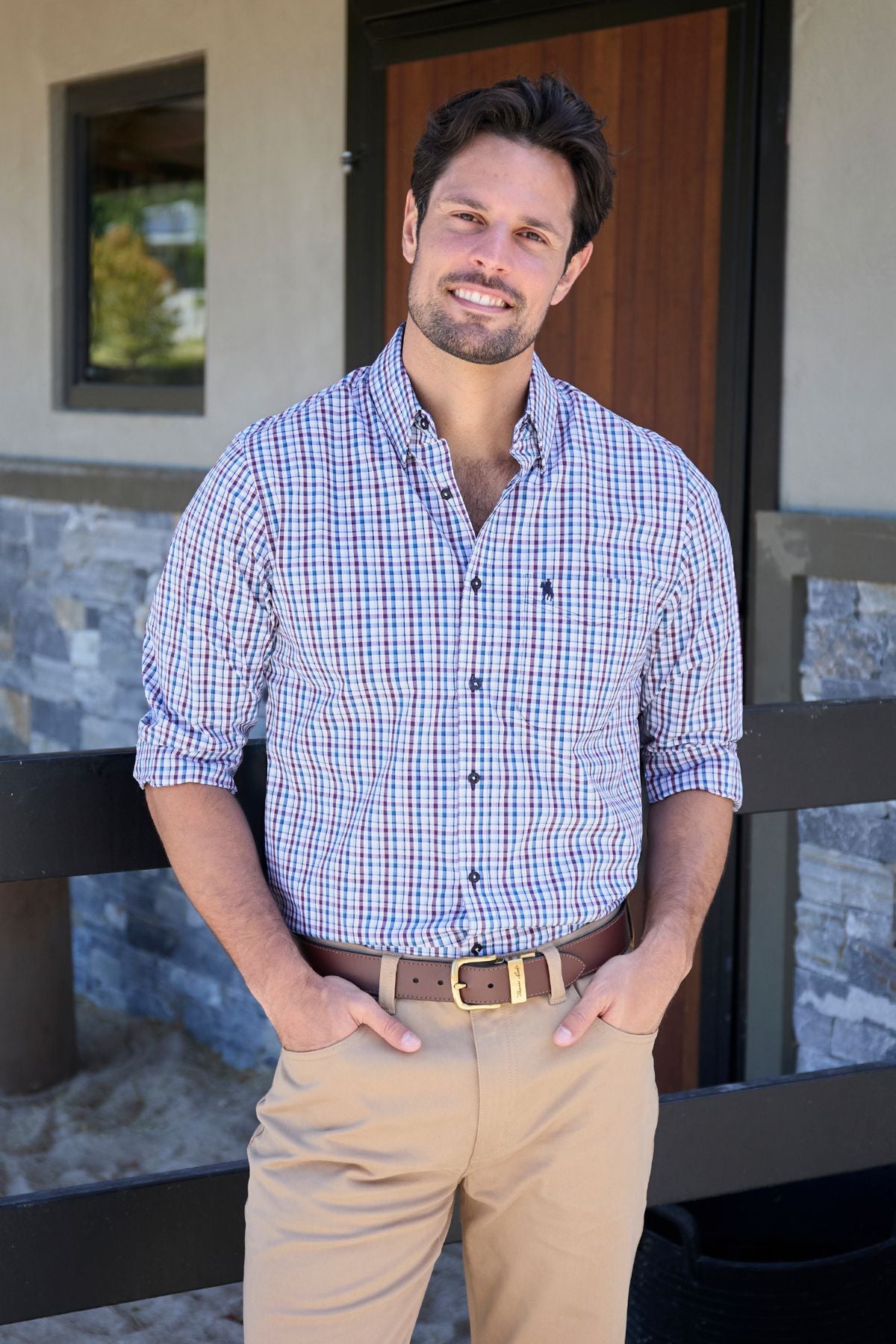 A man with brown hair and a beard smiles outdoors, wearing the Thomas Cook Joseph Shirt in 100% easy-care cotton. His plaid button-down has rolled sleeves, paired with khaki pants, as he stands before a stone and wood building.