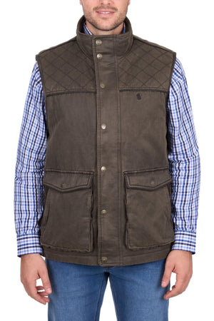 A man wears the Thomas Cook Marcus Vest in brown with snap buttons over a blue checkered shirt and blue jeans, shown from the front and smiling slightly.