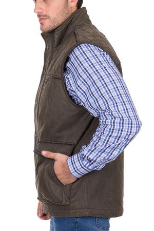 A man in a blue and white plaid shirt and olive green Thomas Cook Marcus Vest with sherpa lining stands in profile, one hand in his pocket, against a white background.