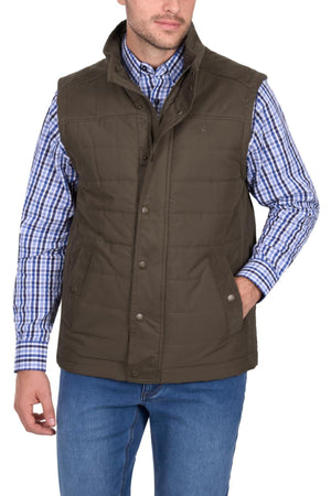 A man wearing the Thomas Cook Matt Vest, a brown quilted vest lined for warmth, over a blue and white plaid shirt and blue jeans, stands with one hand in his pocket.