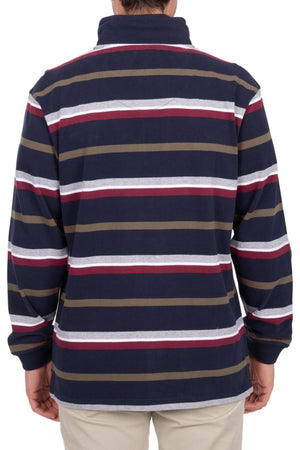 A person is seen from the back wearing the Thomas Cook Nicholas 1/4 Zip Rugby, a heavyweight 100% cotton jersey with navy, white, red, and olive green stripes, paired with light-colored pants.