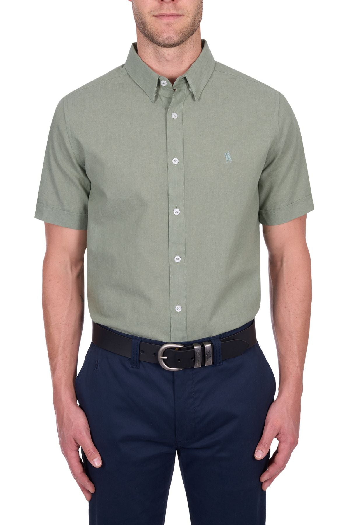 A man wears the Thomas Cook TC Linen Short Sleeve Shirt in light green, tucked into dark blue pants with a black belt, standing against a white background—an effortless summer essential.