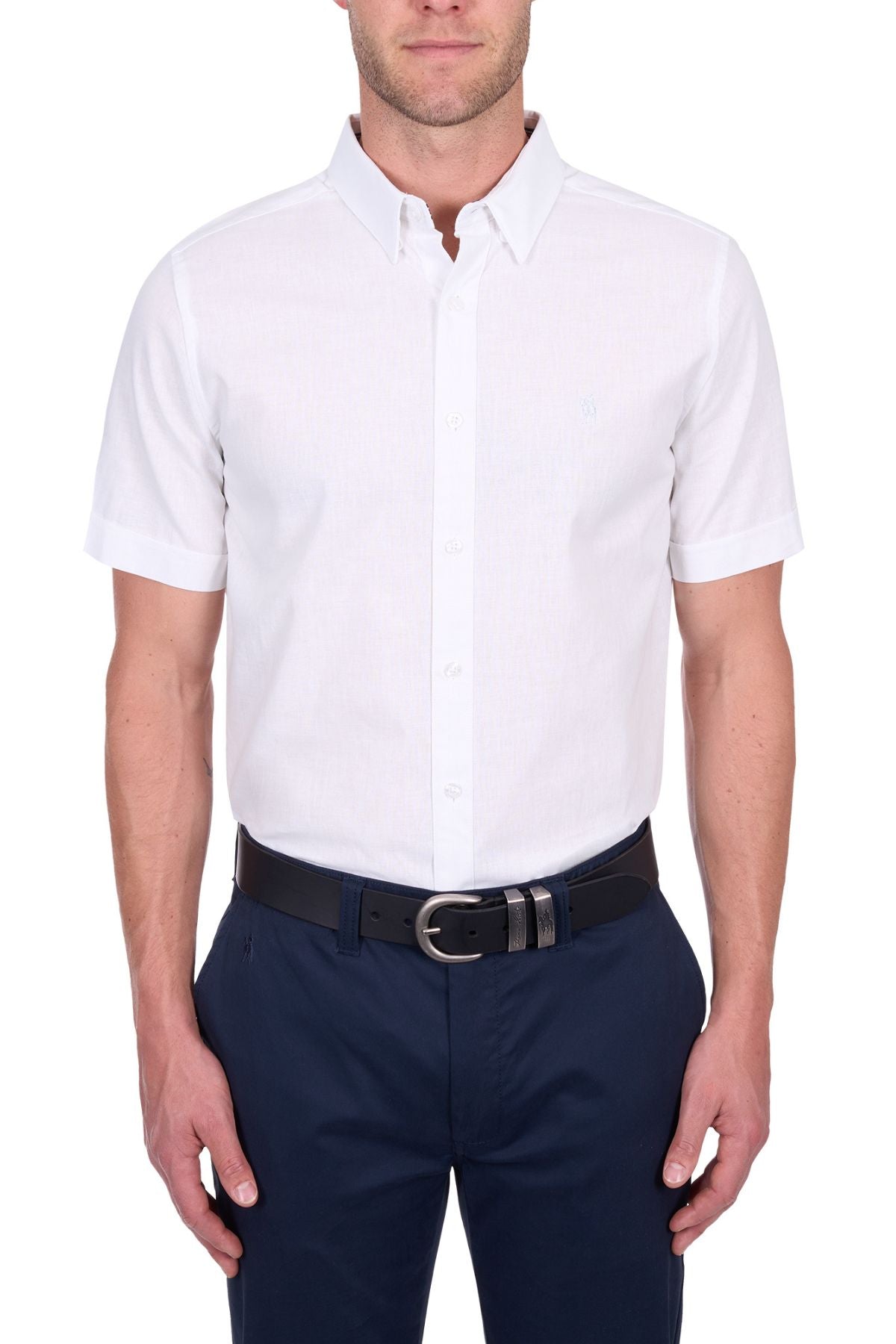 A man stands with arms relaxed at his sides against a white background, wearing the Thomas Cook TC Linen Short Sleeve Shirt in white, paired with navy blue pants and a black belt.