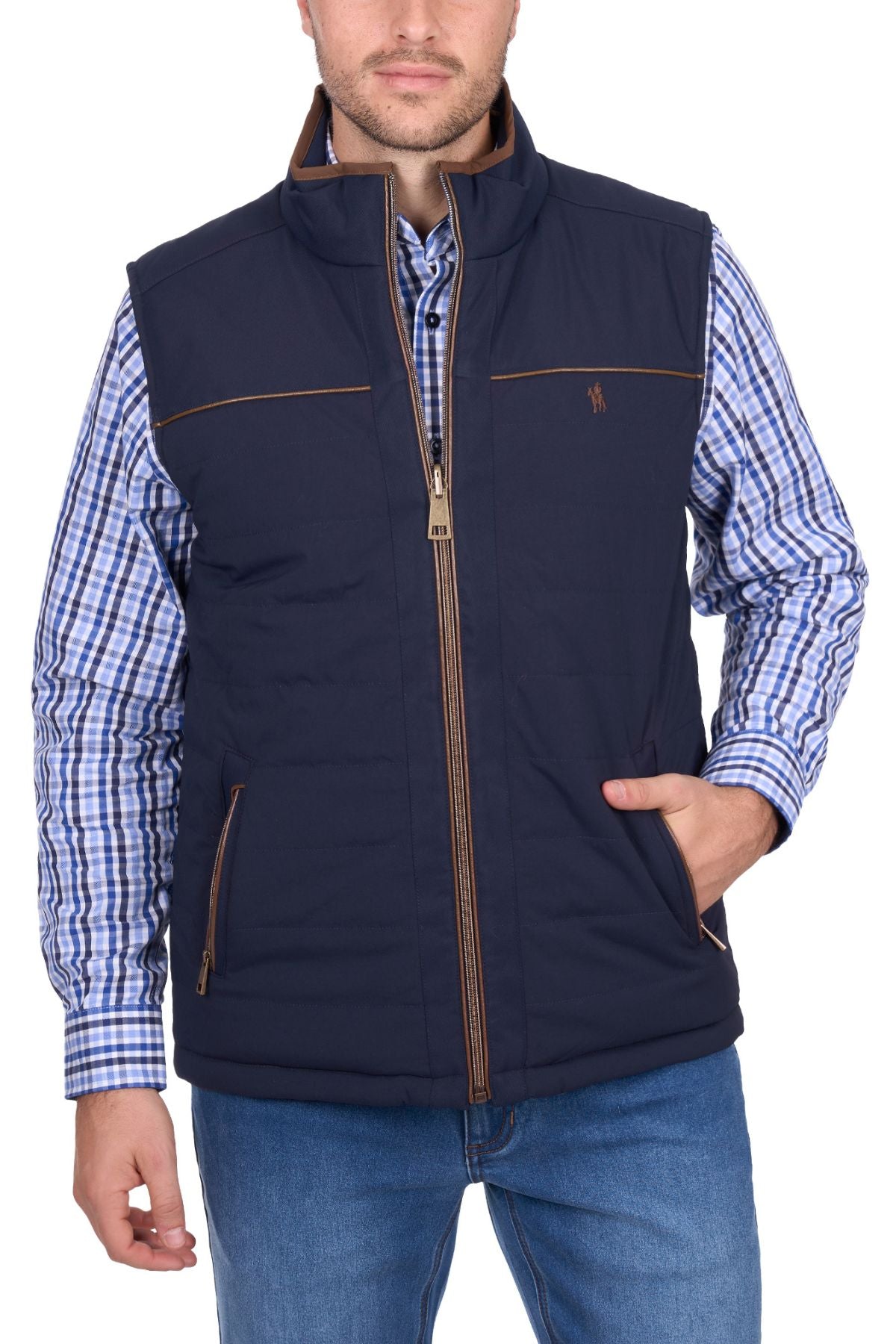 A man stands against a white background wearing the Thomas Cook Toby Revs Vest in blue with brown accents, over a blue checkered shirt and blue jeans, with one hand in his vest pocket.