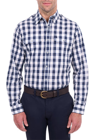 A man stands against a white background, his head partially out of frame, wearing the Thomas Cook Ezra Shirt—a blue and white cotton check workwear shirt—paired with dark trousers and a dark brown belt.