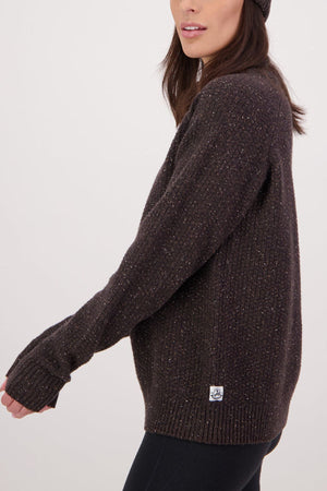 A woman in a dark brown Swanndri Lyttelton Womens Waffle Crew and matching beanie stands sideways against a plain light background, with only her upper body visible as she looks slightly ahead.