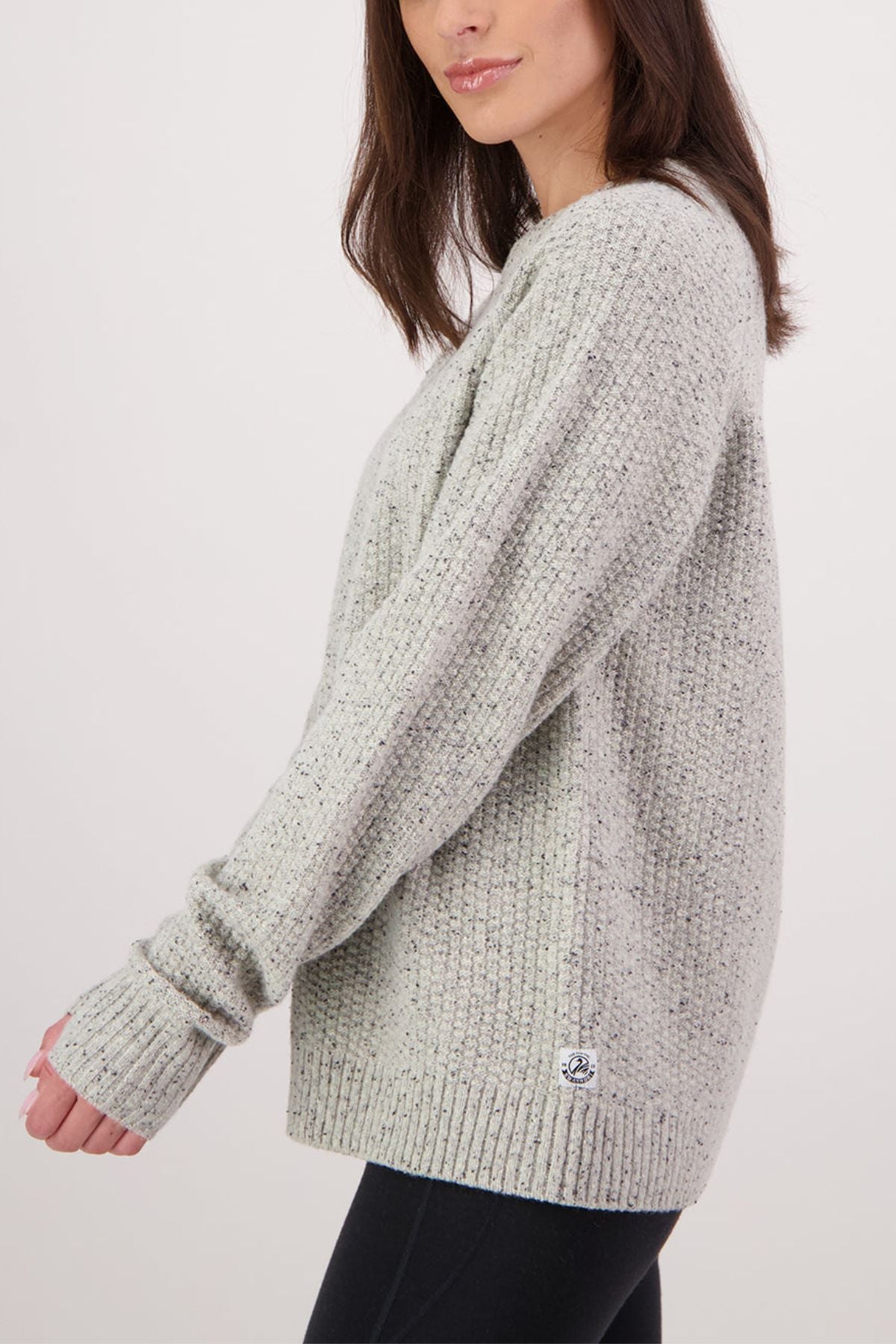 A woman with medium-length brown hair models the Swanndri Lyttelton Womens Waffle Crew, a light gray sweater, with black pants, standing sideways and extending her left arm against a plain white background.