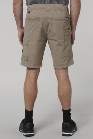 A person faces away from the camera, wearing Hard Yakka X Range Mid Shorts in light brown, with black socks and athletic shoes. The background is plain and light-colored.