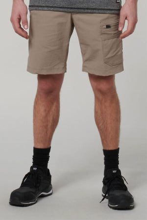 A person stands against a plain light background, wearing Hard Yakka X Range Mid Shorts in beige Cordura nylon, paired with black socks, black sneakers, and a partially visible gray shirt.