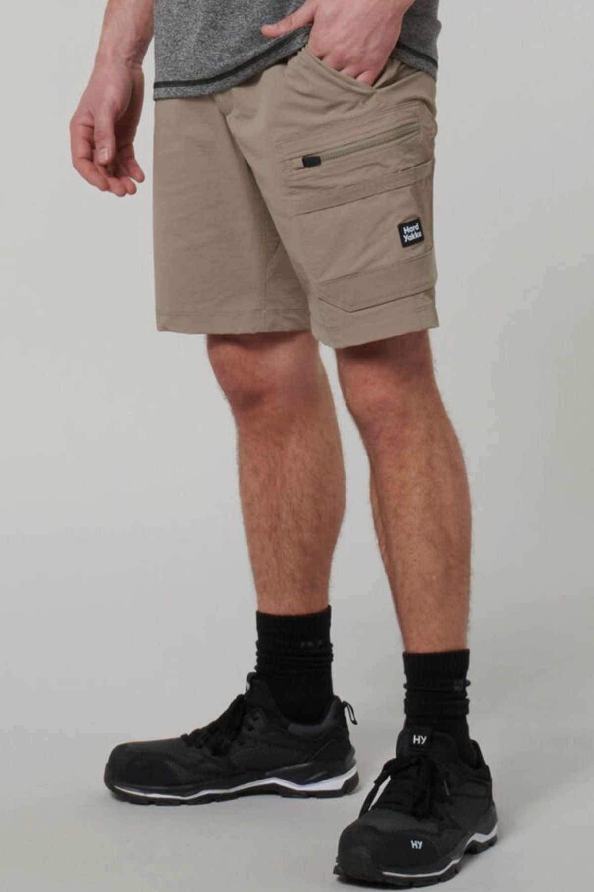 A person wearing Hard Yakka X Range Mid Short cargo shorts in beige Cordura nylon with zipper pockets and a patch label, plus a gray shirt, black socks, and black sneakers, stands against a plain background.