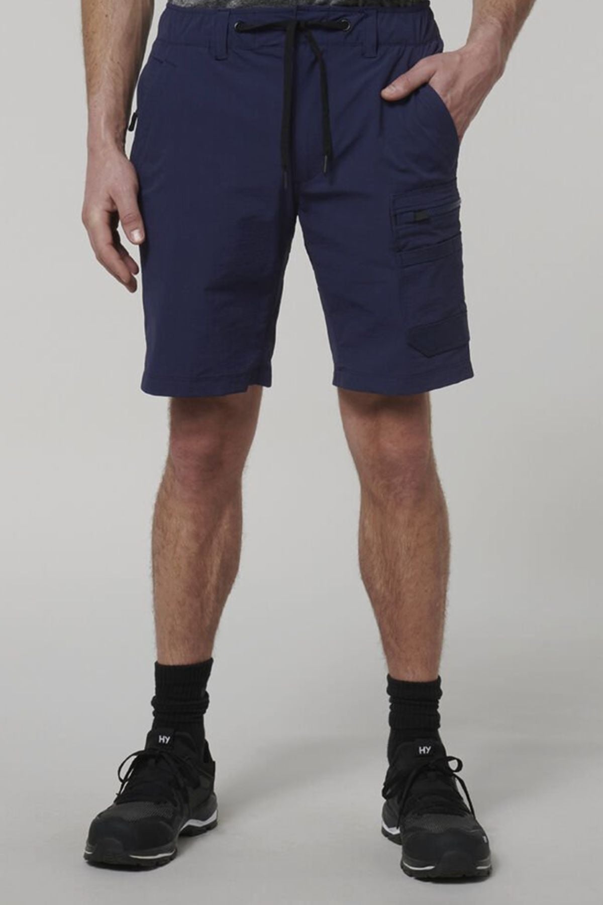 A person models Hard Yakka X Range Mid Shorts in navy blue with utility pockets, styled with black socks and athletic shoes, standing against a light background. The shot highlights the durable work shorts and lower body.