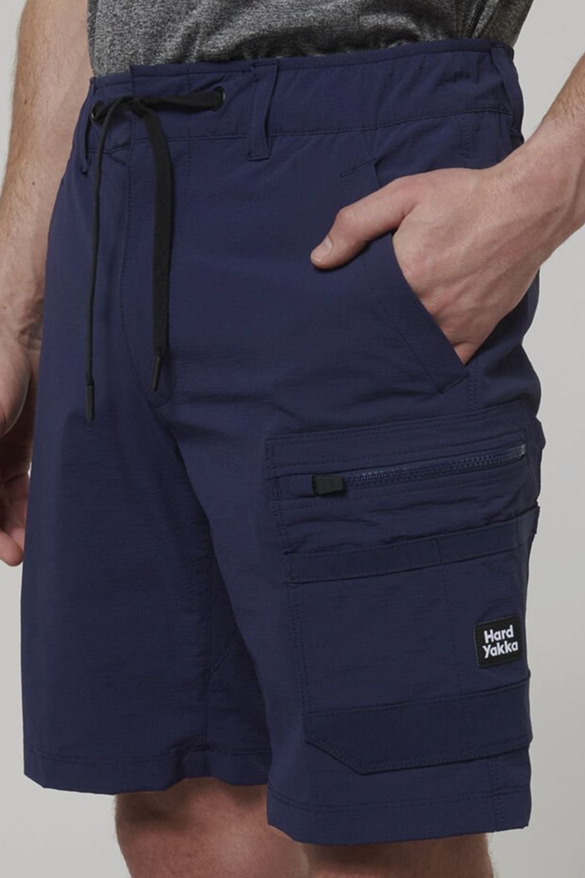 A person wears Hard Yakka X Range Mid Short in navy blue, featuring stretch Cordura fabric, drawstring waist, multiple utility pockets including a zippered and logo pocket, with their hand in one of the front pockets.